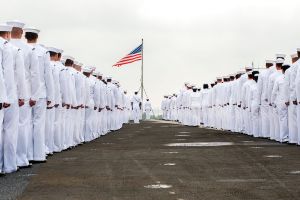 Navy sailors gathered on the deck of a ship in front of a waving American flag.