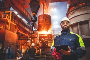 Multiracial man holding tablet looking into the distance standing in front of a metal smelting operation